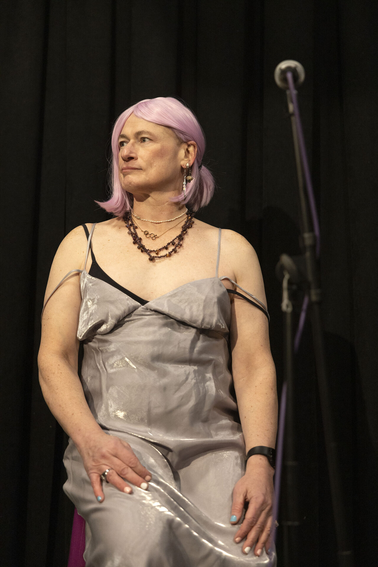 Photograph of Davi Gray. She is a white, trans nonbinary woman of indeterminate age. In this photo she is seated on stage near a microphone; she's wearing a silver gown and pink hair, and is staring offstage to the side.