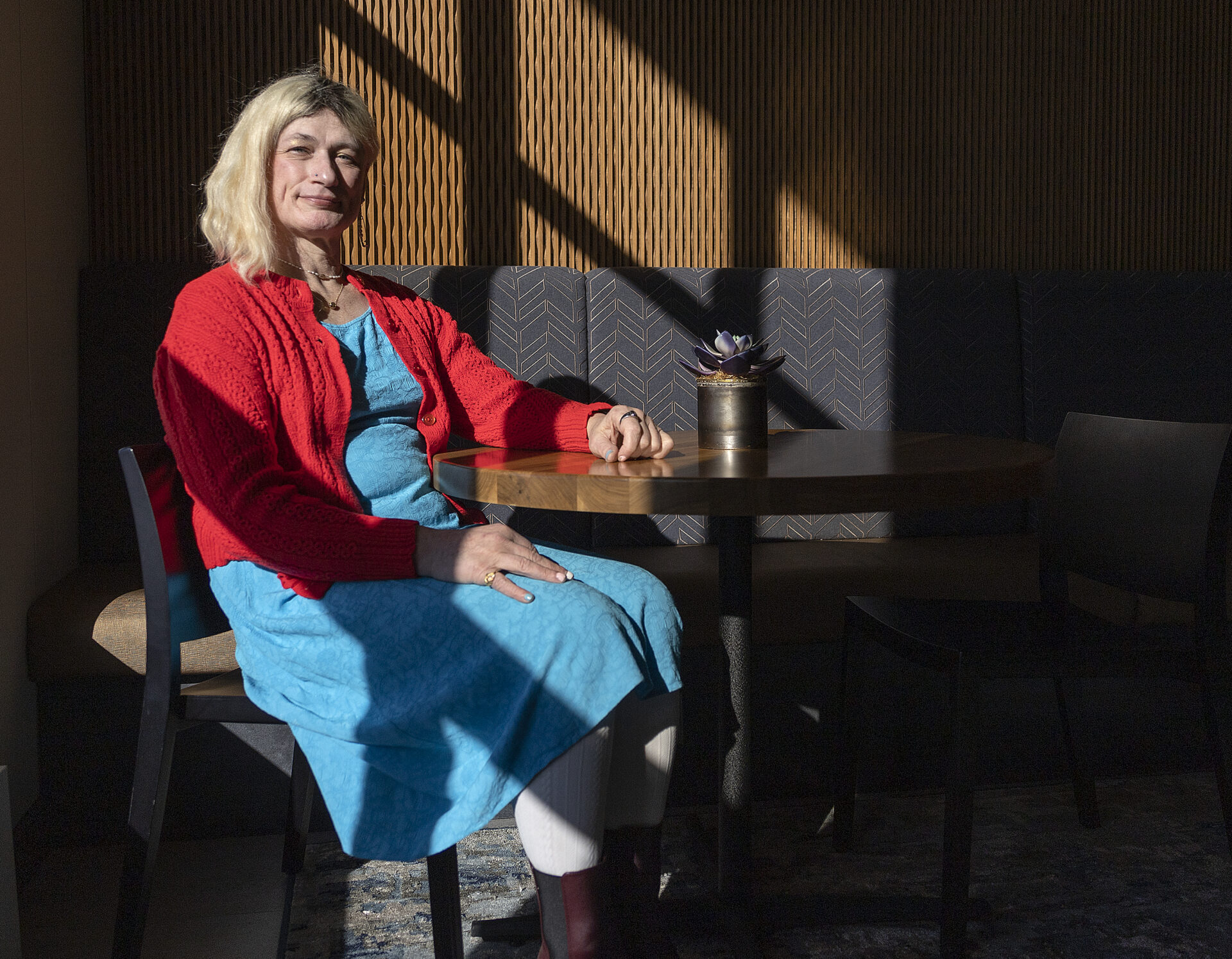 Photo of Davi Gray; she's wearing a bright red cardigan over a baby blue dress with white leggings, and shoulder-length blond hair. She is seated at a table, striped with shadow and sunlight.