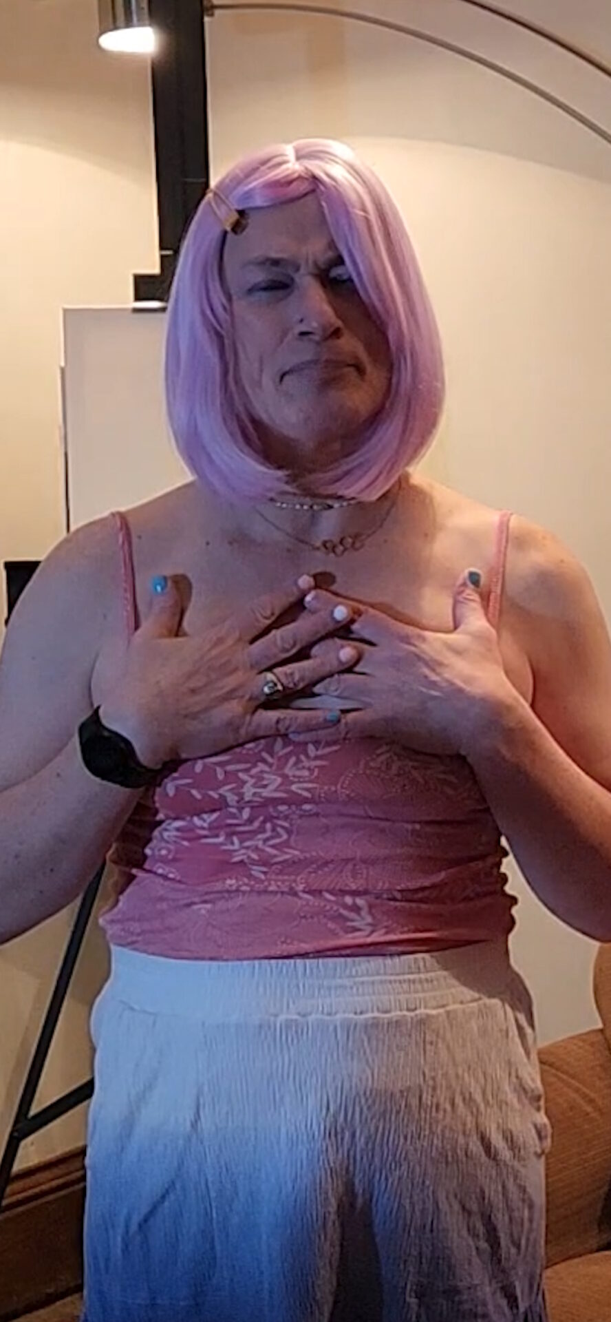 Photo of Davi Gray, wearing pink hair, a flowered pink camisole, and an ombre blue-and-white skirt. Her nails are painted in the colors of the trans pride flag, and she's holding her hands against her chest.